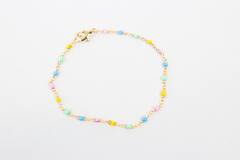 May include: A gold-coloured bracelet with a chain and small, pastel-coloured beads. The bracelet has a lobster clasp. The beads are in pink, yellow, blue, and green.