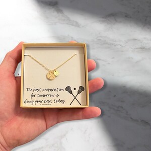 May include: A gold necklace with a lacrosse stick charm and a small round charm in a white box. The box has a quote that reads "The best preparation for tomorrow is doing your best today." - Jackson Brown Jr.