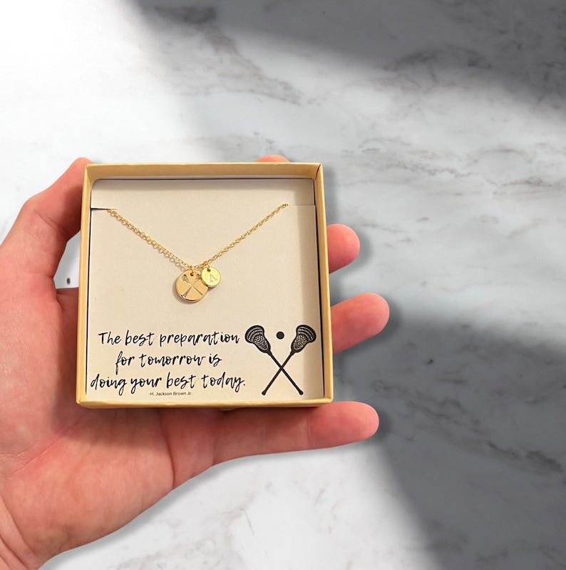 May include: A gold necklace with a lacrosse stick charm and a small round charm in a white box. The box has a quote that reads "The best preparation for tomorrow is doing your best today." - Jackson Brown Jr.