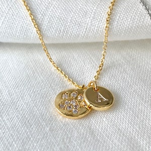 May include: Gold necklace featuring a paw print charm and a letter 'A' charm. The paw print charm is set with small clear stones.