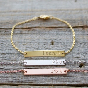 May include: Three gold bar bracelets with personalized engravings. The top bracelet is gold with the name "hannah" engraved on it. The middle bracelet is silver with "p&e" engraved on it. The bottom bracelet is rose gold with "j&hearts;k" engraved on it.