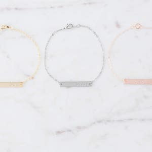 May include: Three delicate gold, silver, and rose gold bar bracelets with a simple, minimalist design. Each bracelet features a thin, rectangular bar with a personalized inscription.