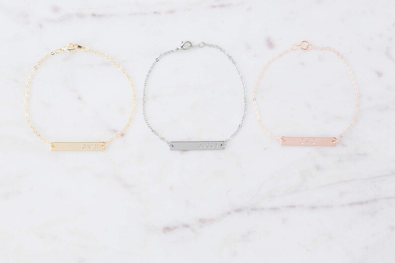 May include: Three delicate gold, silver, and rose gold bar bracelets with a simple, minimalist design. Each bracelet features a thin, rectangular bar with a personalized inscription.