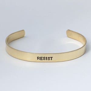 May include: Gold-toned metal bracelet with the word "RESIST" engraved on the front.