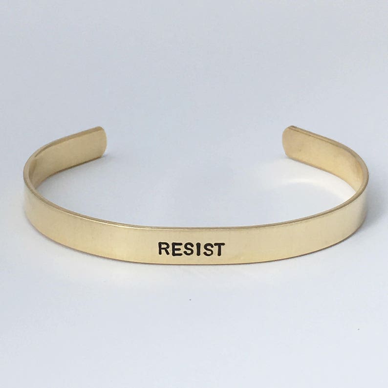 May include: Gold-toned metal bracelet with the word "RESIST" engraved on the front.