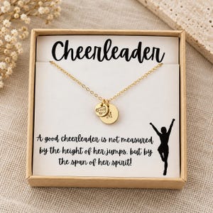 Könnte beinhalten: Eine goldfarbene Halskette mit zwei kleinen runden Anhängern, einer mit einem Megafon-Symbol und der andere mit dem Buchstaben M, präsentiert in einer Schachtel mit dem Text Cheerleader und einem Zitat über Geist sowie einer Silhouette einer springenden Person.