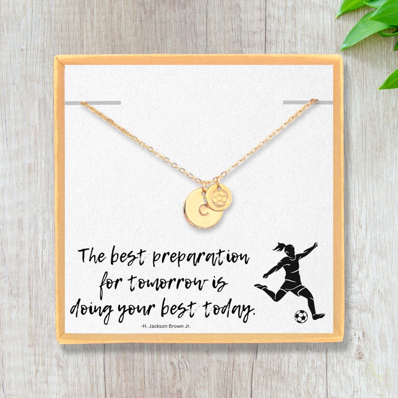 May include: A gold necklace with a soccer ball charm and a letter 'C' charm in a white box with a quote that reads "The best preparation for tomorrow is doing your best today. -H. Jackson Brown Jr."