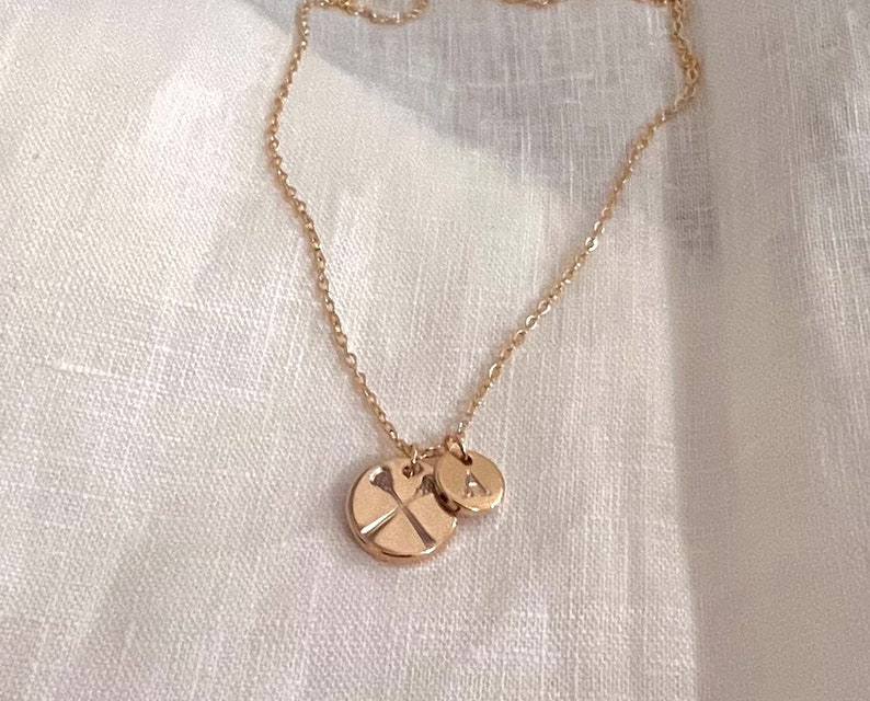 May include: A gold chain necklace with two charms. One charm is a round disc with a cross design, and the other charm is a round disc with the letter "A" engraved on it.