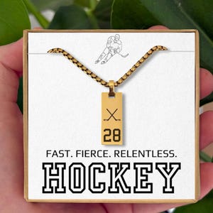 May include: Gold-colored hockey necklace with a rectangular pendant featuring crossed hockey sticks and the number 28. The card reads "FAST. FIERCE. RELENTLESS. HOCKEY."