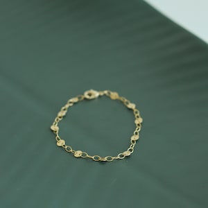 May include: A delicate gold bracelet featuring a chain of small, flat, circular charms. The bracelet is secured with a lobster clasp. The jewelry is displayed on a dark green surface.