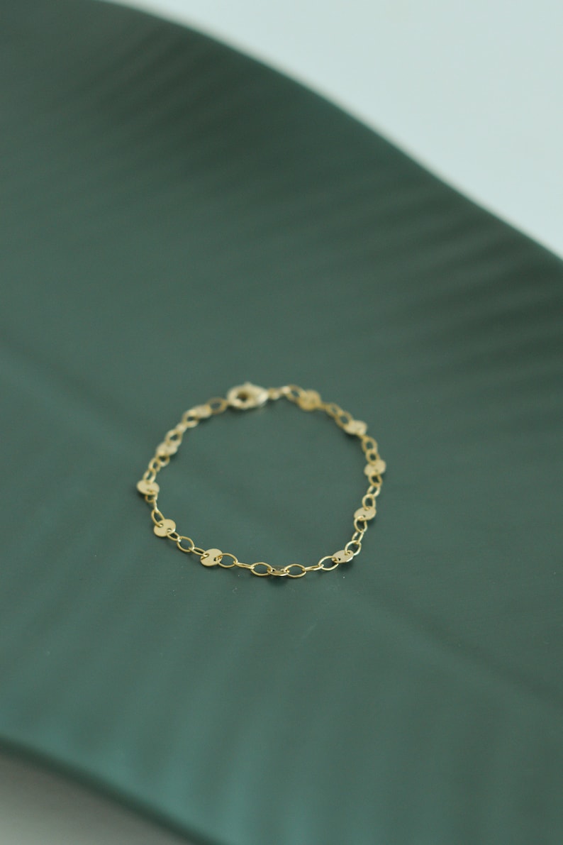 May include: A delicate gold bracelet featuring a chain of small, flat, circular charms. The bracelet is secured with a lobster clasp. The jewelry is displayed on a dark green surface.