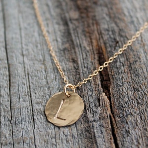 Puede incluir: Un collar de cadena de oro con un colgante redondo que presenta la letra "L" en relieve. El colgante tiene una textura martillada.