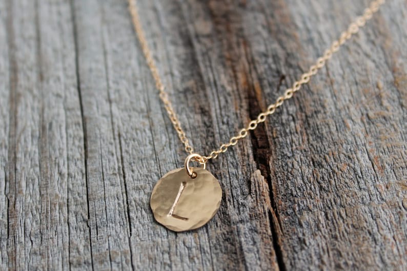 Puede incluir: Un collar de cadena de oro con un colgante redondo que presenta la letra "L" en relieve. El colgante tiene una textura martillada.