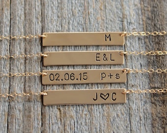 Op de afbeelding: Vier gouden bar kettingen met verschillende gravures. De bovenste ketting heeft de letter "M" gegraveerd op de bar. De tweede ketting heeft "E & L" gegraveerd op de bar. De derde ketting heeft "02.06.15" en "p + s" gegraveerd op de bar. De onderste ketting heeft "J ♥ C" gegraveerd op de bar.