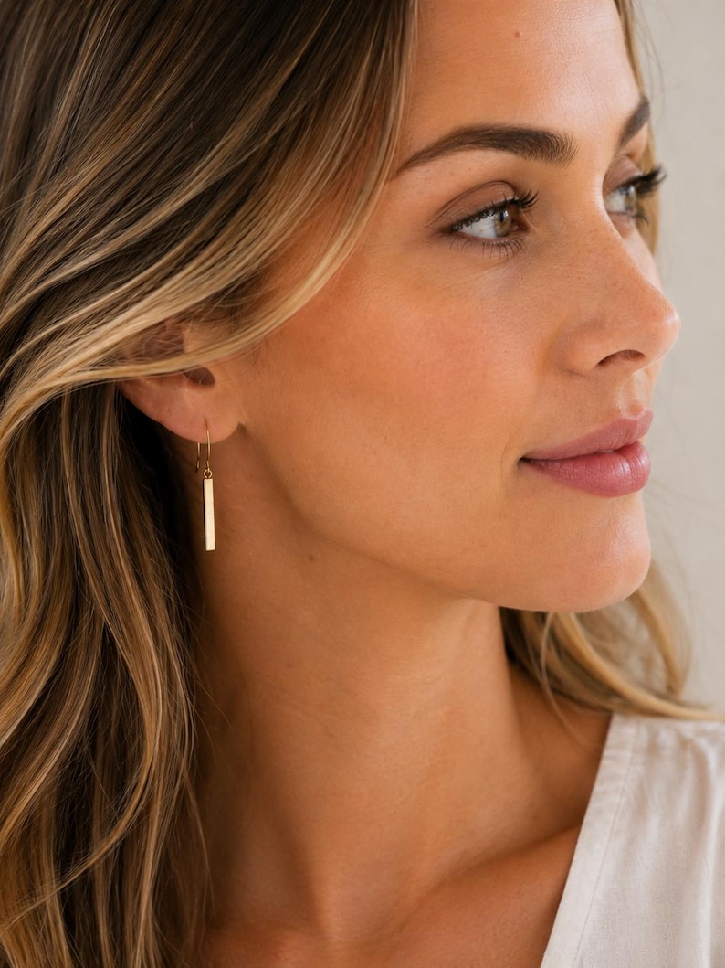 May include: A gold-toned drop earring featuring a thin, rectangular bar pendant suspended from a simple curved wire hook. The metal surface has a smooth, polished finish that reflects light, creating a clean and straightforward geometric design.