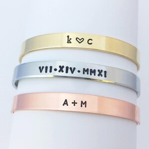 May include: Three stacked bracelets with different engravings. The top bracelet is gold and says "k &hearts; c". The middle bracelet is silver and says "VII.XIV.MMXI". The bottom bracelet is copper and says "A + M".