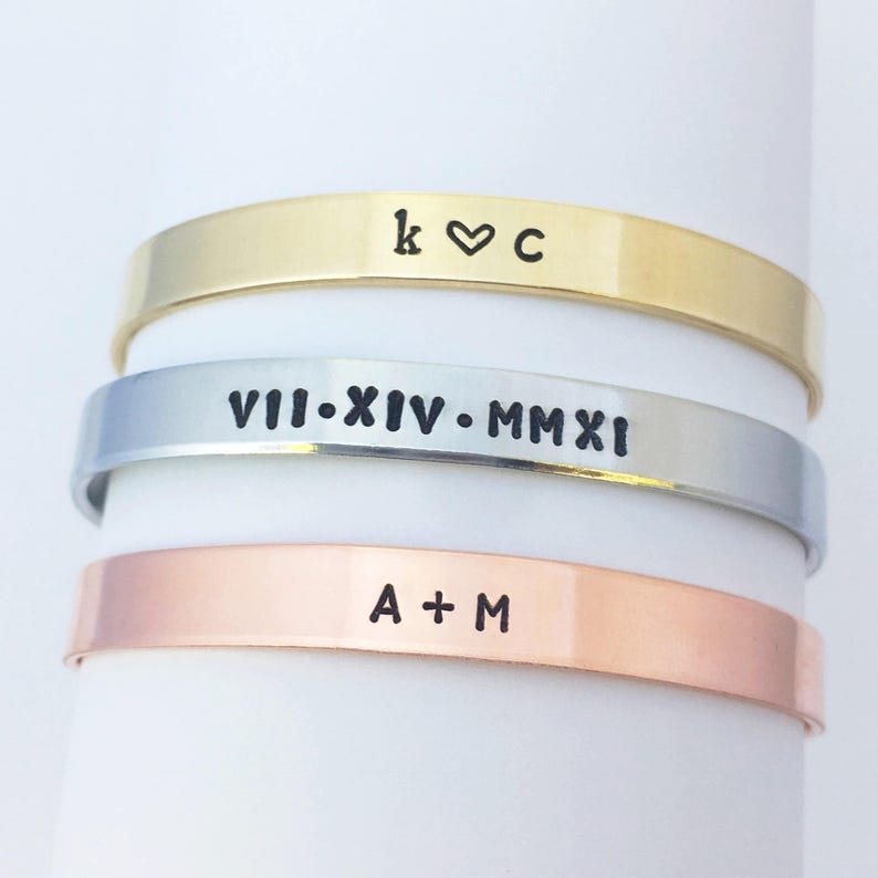 May include: Three stacked bracelets with different engravings. The top bracelet is gold and says "k &hearts; c". The middle bracelet is silver and says "VII.XIV.MMXI". The bottom bracelet is copper and says "A + M".
