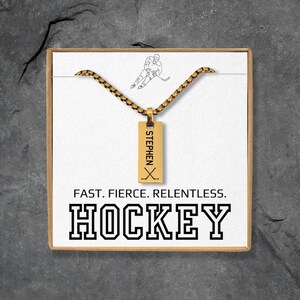 May include: A gold-colored hockey pendant necklace with the name "STEPHEN" and hockey sticks. The necklace is presented in a gift box with the words "FAST. FIERCE. RELENTLESS. HOCKEY."