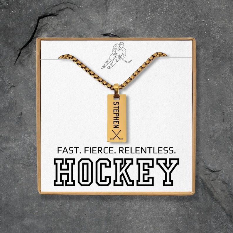 May include: A gold-colored hockey pendant necklace with the name "STEPHEN" and hockey sticks. The necklace is presented in a gift box with the words "FAST. FIERCE. RELENTLESS. HOCKEY."