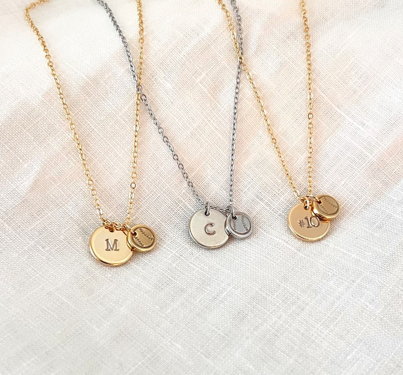 May include: Three gold necklaces with a baseball charm and a letter charm. The charms are engraved with the letters "M", "C", and the number "10".
