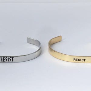 May include: Two simple silver and gold bracelets with the word "RESIST" engraved on them.