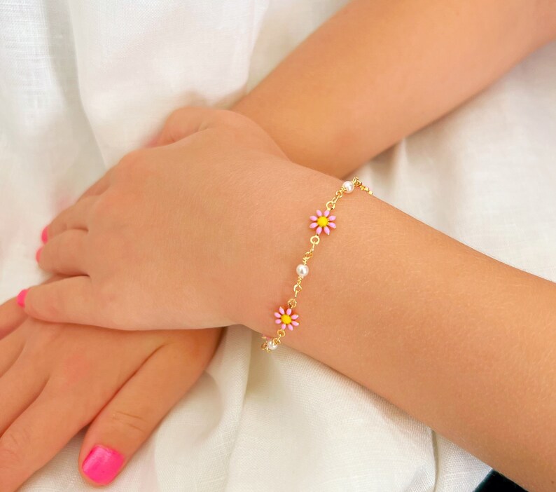 May include: A gold chain bracelet with pink and yellow daisy flower charms and small white pearls. The bracelet is on a wrist with pink nail polish.