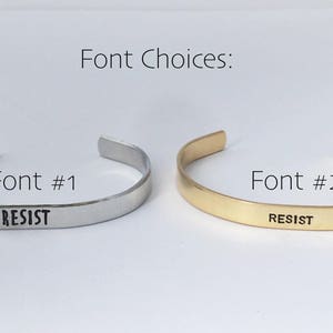 May include: Two silver and gold bracelets with the word "RESIST" engraved on them. The bracelets are shown side-by-side, with the silver bracelet on the left and the gold bracelet on the right. The bracelets are both simple and elegant, with a smooth finish. The word "RESIST" is engraved in a simple, sans-serif font. The bracelets are a great way to show your support for a cause or to make a statement.