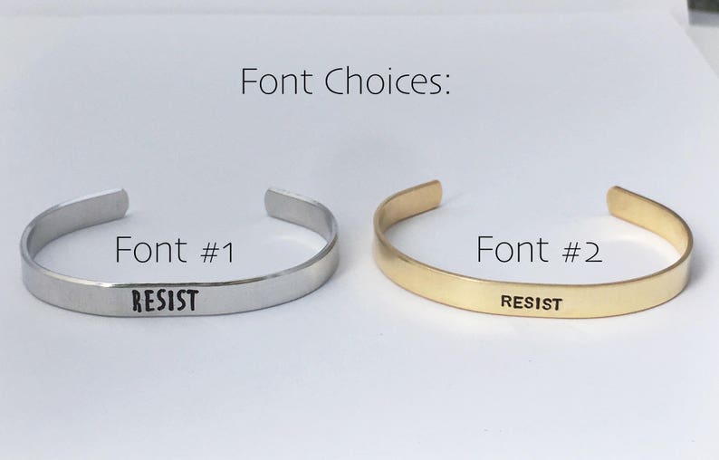 May include: Two silver and gold bracelets with the word "RESIST" engraved on them. The bracelets are shown side-by-side, with the silver bracelet on the left and the gold bracelet on the right. The bracelets are both simple and elegant, with a smooth finish. The word "RESIST" is engraved in a simple, sans-serif font. The bracelets are a great way to show your support for a cause or to make a statement.