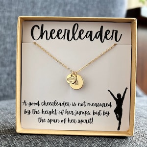 May include: A gold necklace with a charm that says "CHEER" and a small round charm. The necklace is in a white box with the words "Cheerleader" and a quote: "A good cheerleader is not measured by the height of her jumps, but by the span of her spirit!"