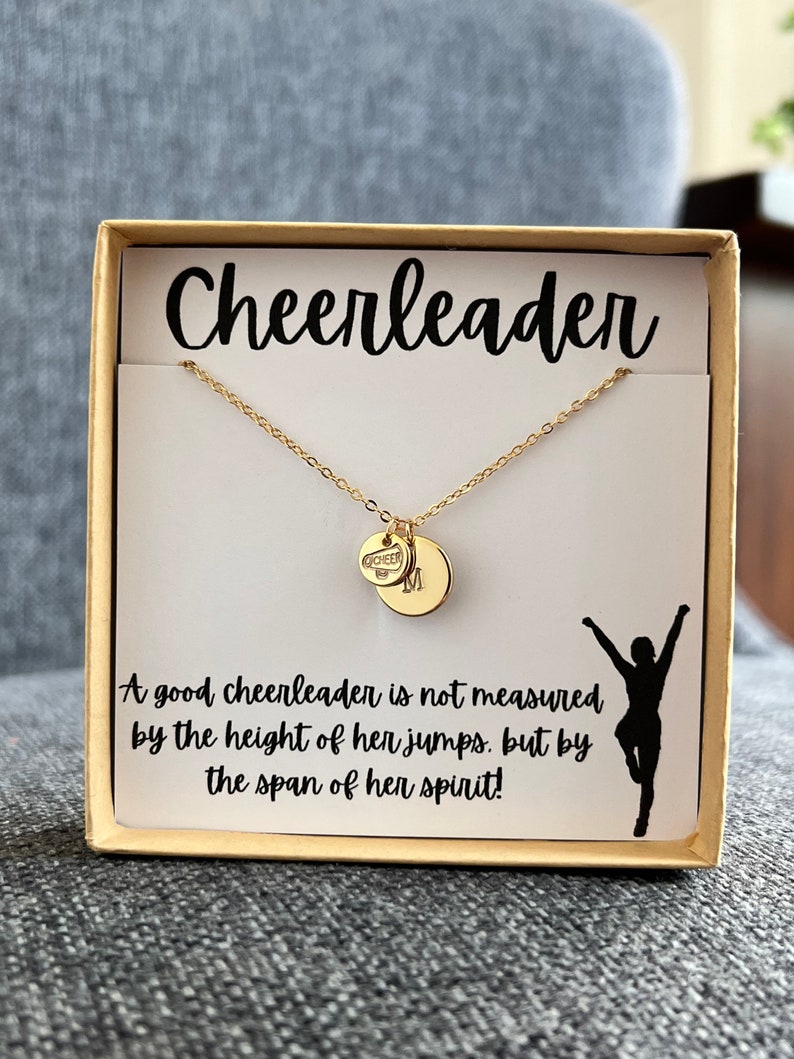 May include: A gold necklace with a charm that says "CHEER" and a small round charm. The necklace is in a white box with the words "Cheerleader" and a quote: "A good cheerleader is not measured by the height of her jumps, but by the span of her spirit!"