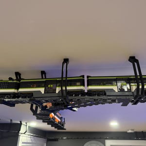 May include: A model train set suspended from a ceiling using black mounting brackets. The train features a green, white, and black color scheme with transparent windows and detailed wheel assemblies, positioned on a track system attached overhead.