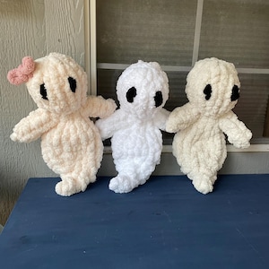 May include: Three handmade crochet ghost dolls in varying shades of white and cream. One ghost has a pink bow. Each ghost has black oval eyes and small arms. The dolls are displayed on a blue surface.