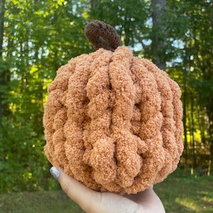 May include: A round, crocheted pumpkin made from soft, textured peach-colored yarn. The pumpkin features a small, dark brown crocheted stem at the top, with a ribbed, plush appearance created by the thick, looped yarn construction.