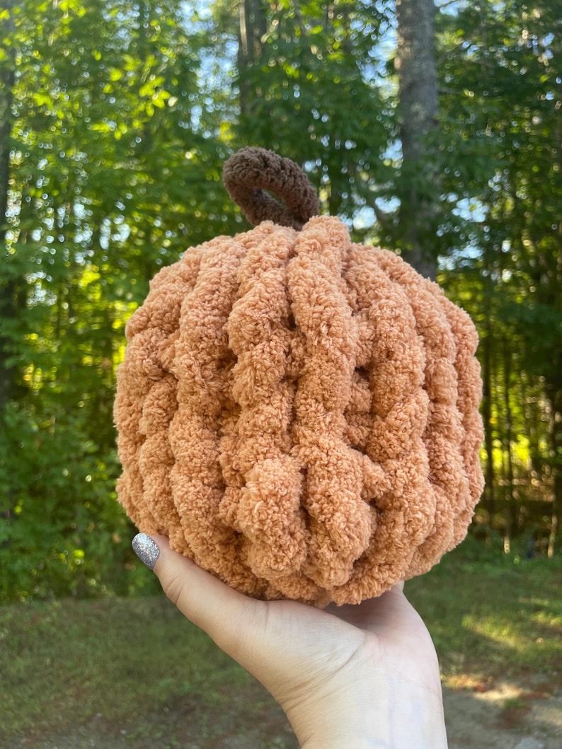 May include: A round, crocheted pumpkin made from soft, textured peach-colored yarn. The pumpkin features a small, dark brown crocheted stem at the top, with a ribbed, plush appearance created by the thick, looped yarn construction.