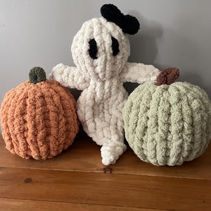 May include: A set of three handmade crochet plush items consisting of a white ghost with black eyes and a black bow, an orange pumpkin with a dark green stem, and a light green pumpkin with a brown stem, all made from soft, chunky yarn.