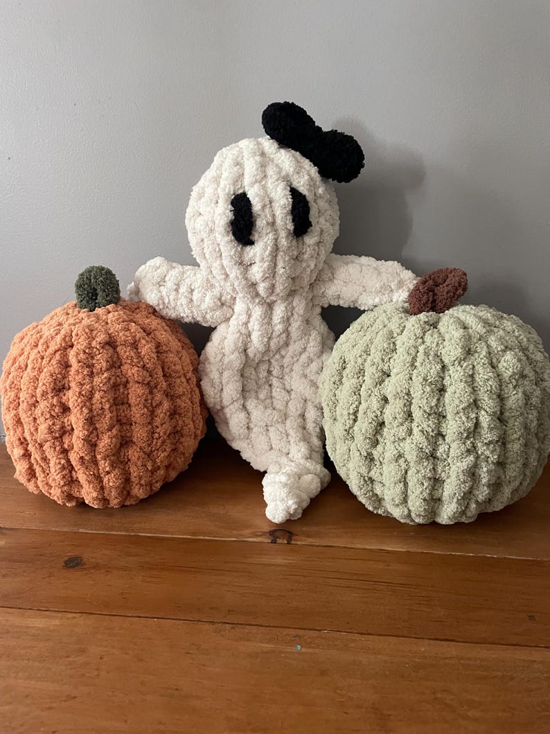 May include: A set of three handmade crochet plush items consisting of a white ghost with black eyes and a black bow, an orange pumpkin with a dark green stem, and a light green pumpkin with a brown stem, all made from soft, chunky yarn.