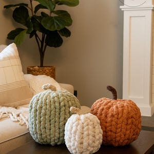 May include: Three handmade chunky knit pumpkins in sage green, cream, and burnt orange. Each pumpkin features a textured, soft yarn construction with a small, distinct stem on top. The pumpkins are arranged together on a dark wooden surface.
