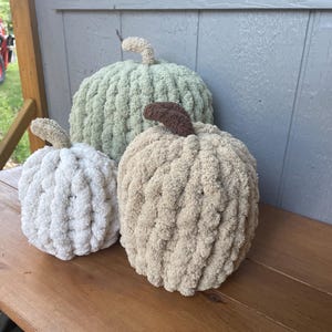 May include: Three handmade pumpkins crafted from chunky, soft yarn in varying sizes. The largest pumpkin is sage green, the medium one is beige with a dark brown stem, and the smallest is white. Each pumpkin features a textured, looped knit surface.