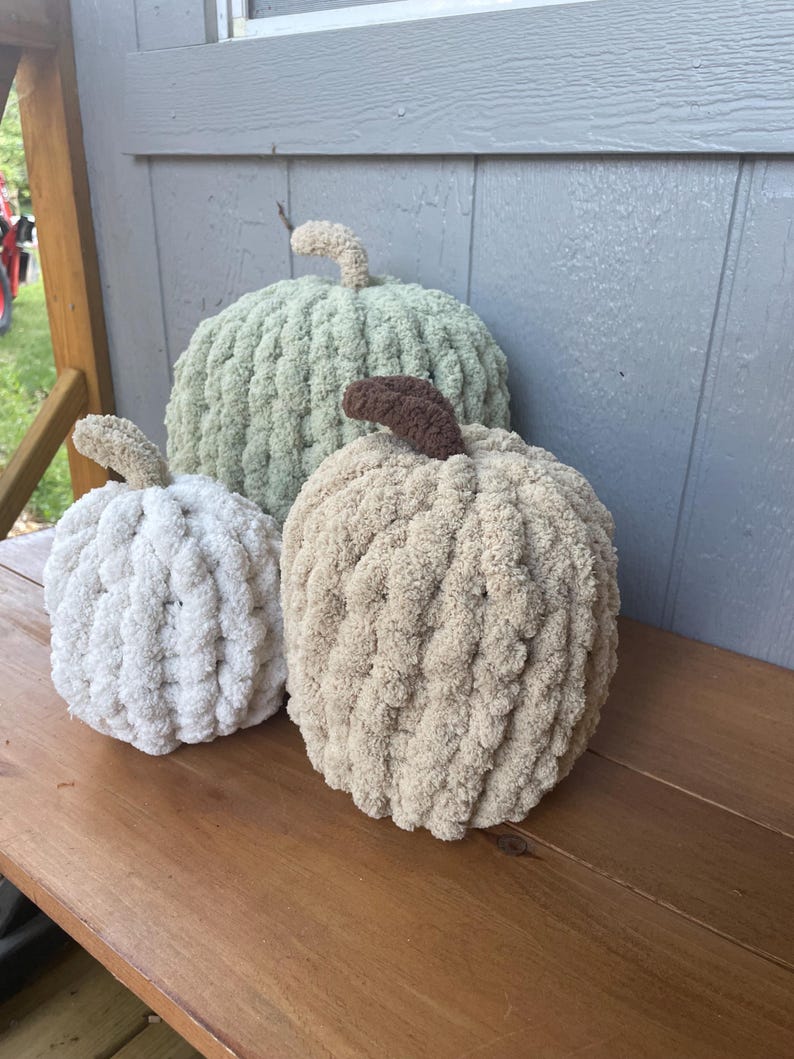 May include: Three handmade pumpkins crafted from chunky, soft yarn in varying sizes. The largest pumpkin is sage green, the medium one is beige with a dark brown stem, and the smallest is white. Each pumpkin features a textured, looped knit surface.