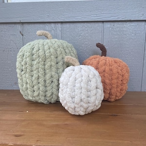 May include: Three handmade pumpkins crafted from chunky, soft yarn. The largest pumpkin is sage green with a tan stem, the medium one is burnt orange with a dark brown stem, and the smallest is cream-colored with a tan stem.