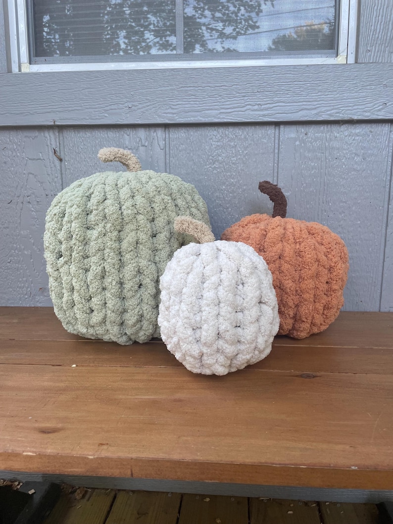 May include: Three handmade pumpkins crafted from chunky, soft yarn. The largest pumpkin is sage green with a tan stem, the medium one is burnt orange with a dark brown stem, and the smallest is cream-colored with a tan stem.