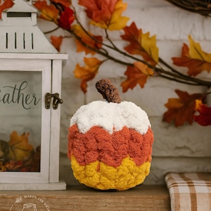 May include: A plush, crocheted pumpkin decoration featuring horizontal color-blocked stripes of white, orange, and yellow. The pumpkin has a textured, bumpy surface and a brown, gnarled stem on top, mimicking the appearance of a piece of candy corn.