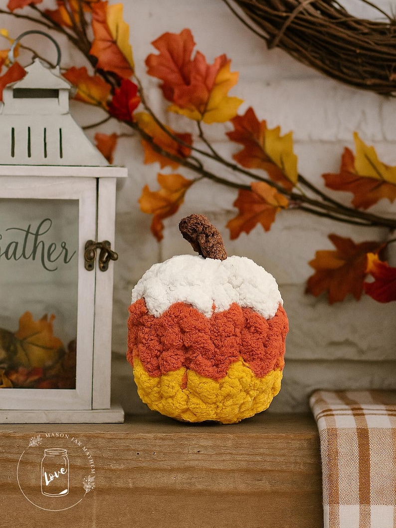 May include: A plush, crocheted pumpkin decoration featuring horizontal color-blocked stripes of white, orange, and yellow. The pumpkin has a textured, bumpy surface and a brown, gnarled stem on top, mimicking the appearance of a piece of candy corn.