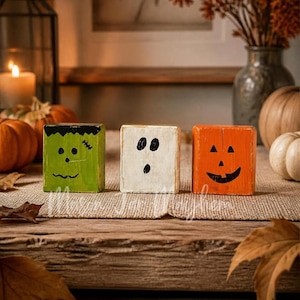 May include: Three rectangular wooden blocks painted with Halloween characters. The left block is green with a black hair and stitch design. The center block is white with a ghost face. The right block is orange with a jack-o'-lantern face.