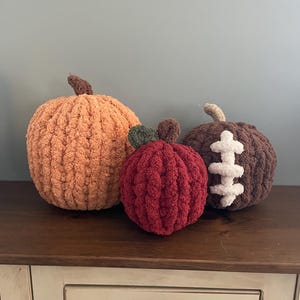 May include: Three plush, crocheted decorative items: a large orange pumpkin with a brown stem, a smaller red apple with a green leaf and brown stem, and a dark brown pumpkin featuring white, stitched-on laces resembling a football design.