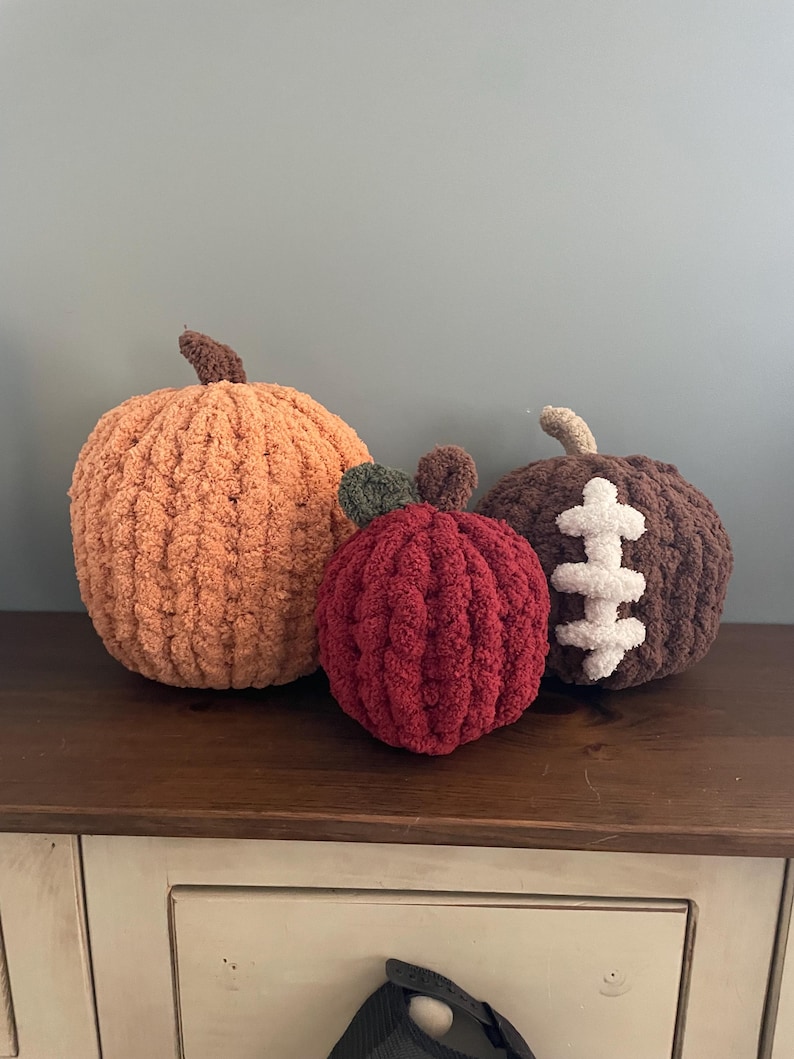 May include: Three plush, crocheted decorative items: a large orange pumpkin with a brown stem, a smaller red apple with a green leaf and brown stem, and a dark brown pumpkin featuring white, stitched-on laces resembling a football design.