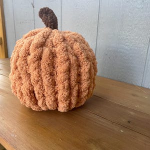 May include: A round, plush pumpkin crafted from soft, peach-colored yarn with a ribbed texture. The pumpkin features a small, dark brown crocheted stem protruding from the top center, creating a rounded, stuffed form with a fuzzy, tactile appearance.