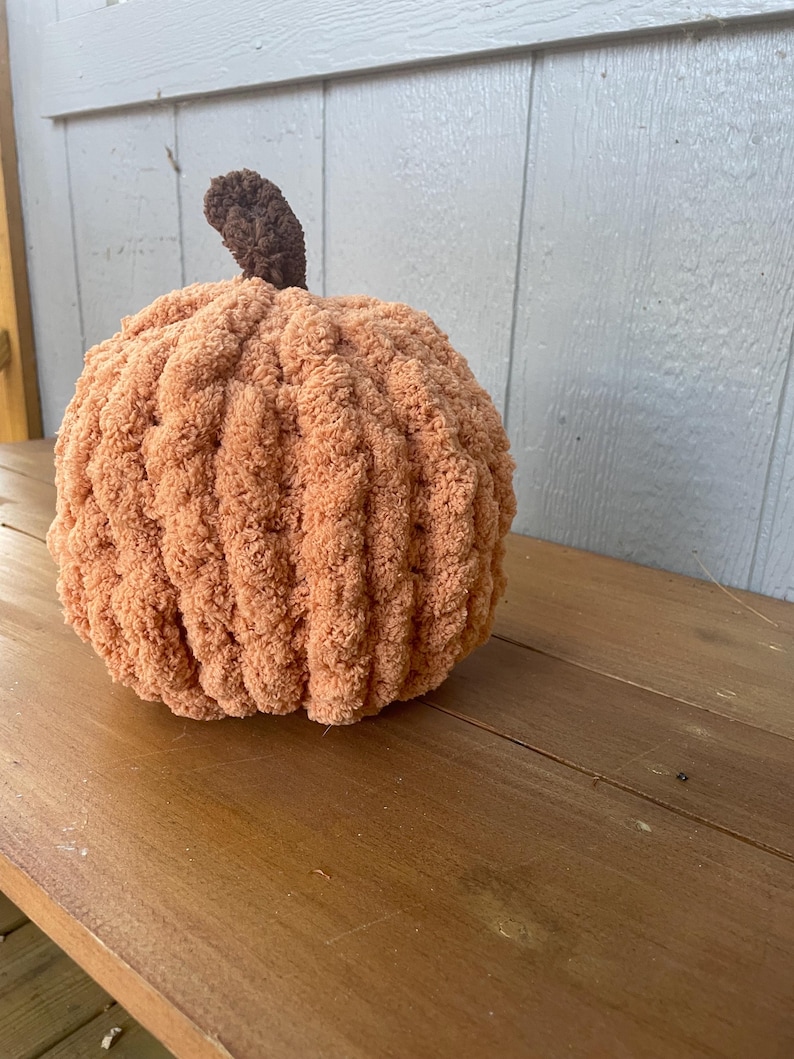 May include: A round, plush pumpkin crafted from soft, peach-colored yarn with a ribbed texture. The pumpkin features a small, dark brown crocheted stem protruding from the top center, creating a rounded, stuffed form with a fuzzy, tactile appearance.