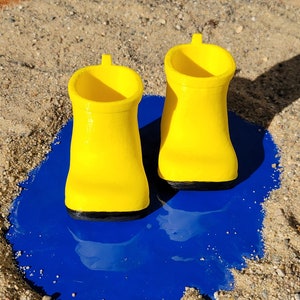 May include: Two yellow rain boots with black soles sit in a blue puddle on a sandy surface.