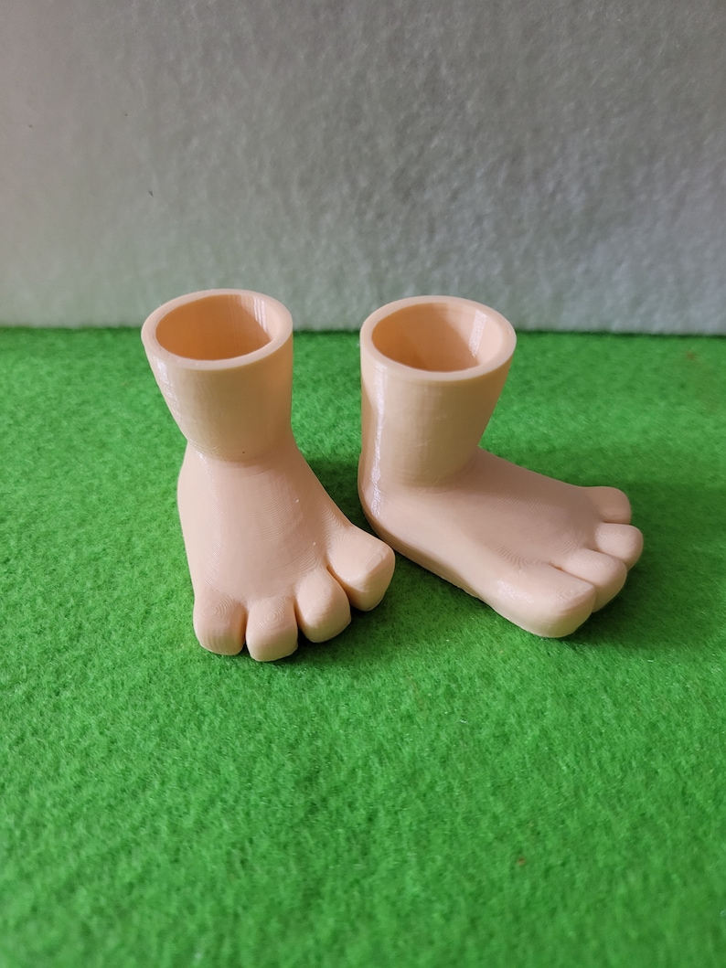 K&ouml;nnte beinhalten: Ein Paar 3D-gedruckte, hellfarbene, pupfengro&szlig;e F&uuml;&szlig;e mit Zehen. Die F&uuml;&szlig;e befinden sich auf einer gr&uuml;nen Oberfl&auml;che.